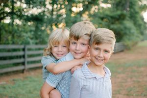 Three blonde brothers in striped shirts smile and hug each other in front of a wooden fence outdoors.