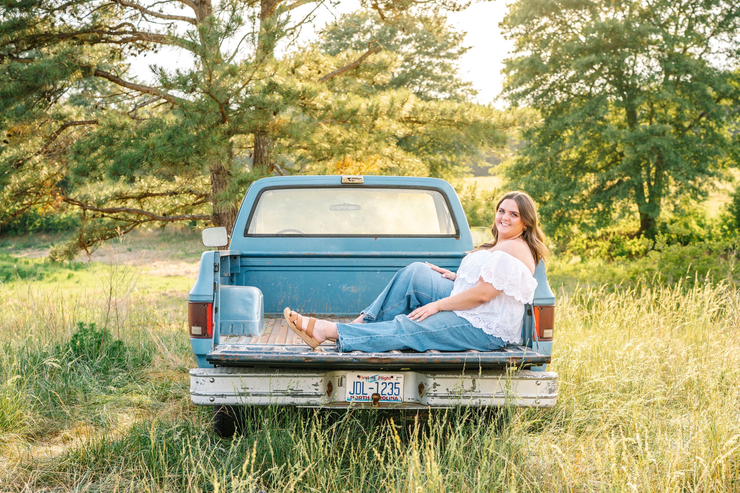 Young woman in white top and jeans sitting in the bed of a vintage teal pickup truck in a sunny field.