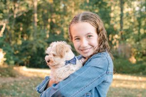 A smiling young girl holds an adorable fluffy puppy wearing a bow tie in a sunny park.