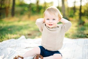 Adorable blonde toddler boy sitting on a blanket outdoors with hands behind head smiling in sunlight.