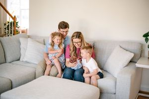 Happy family of five sitting together on a gray couch in a bright living room with a newborn.