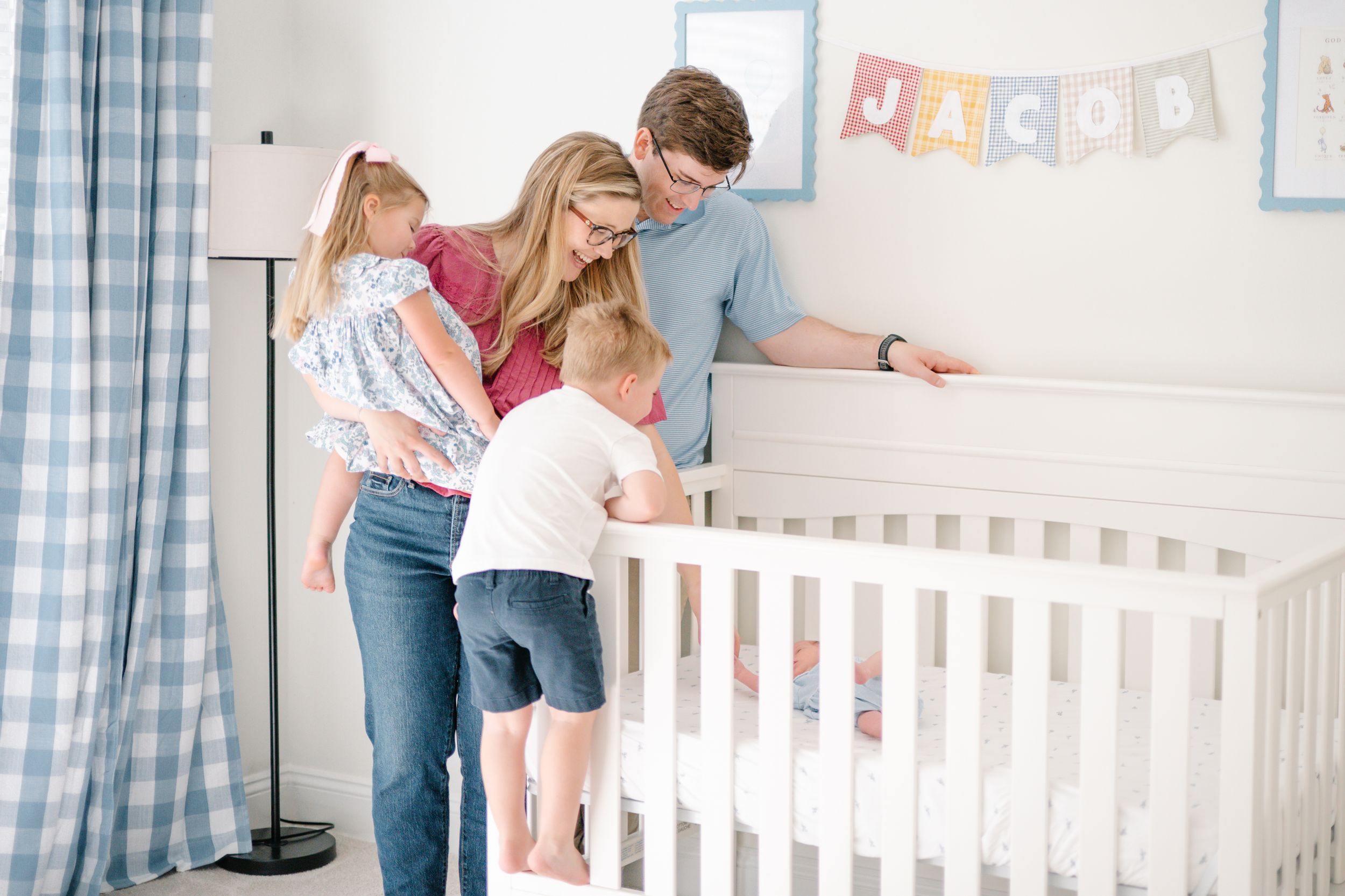 Parents and two young children looking into a white crib in a baby boy nursery decorated in blue.
