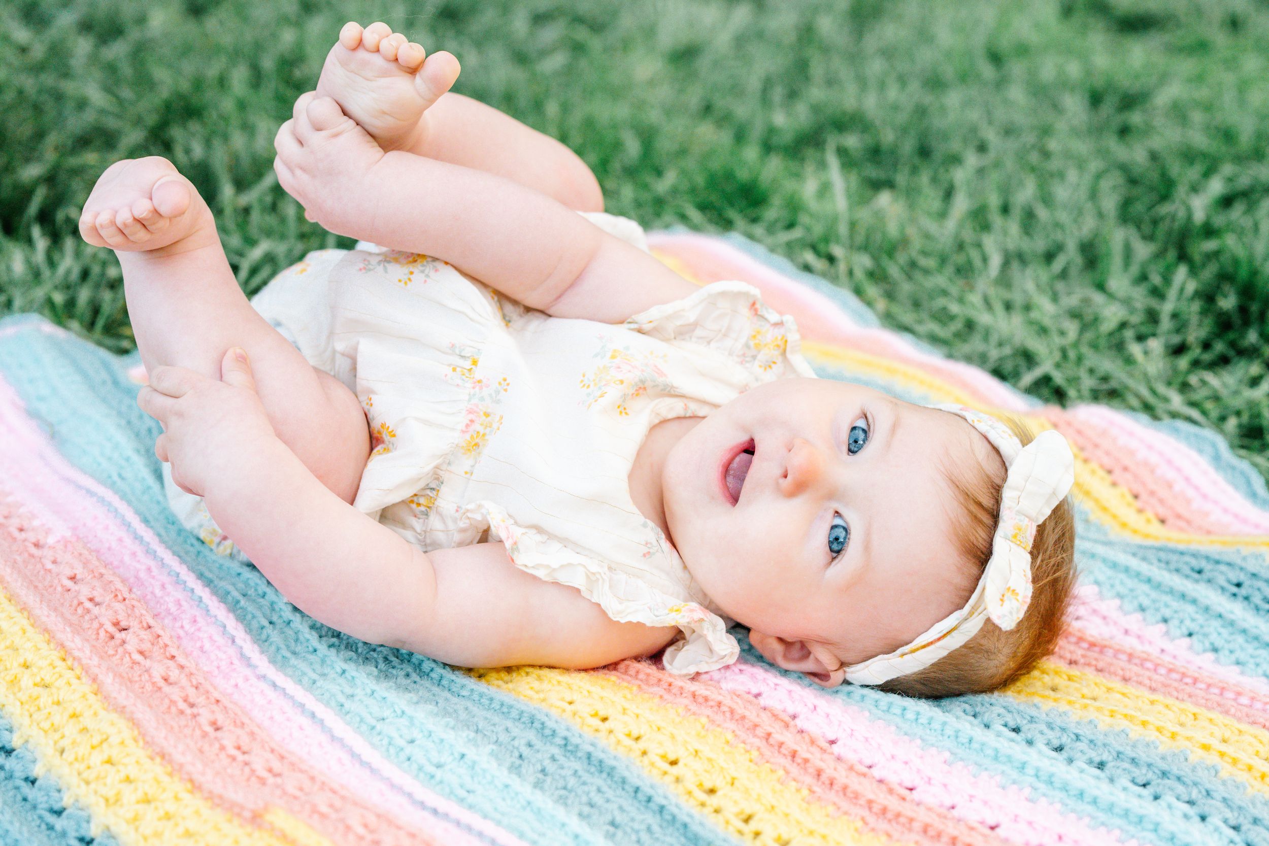 Happy baby girl in white romper lying on a colorful crocheted rainbow blanket on green grass.