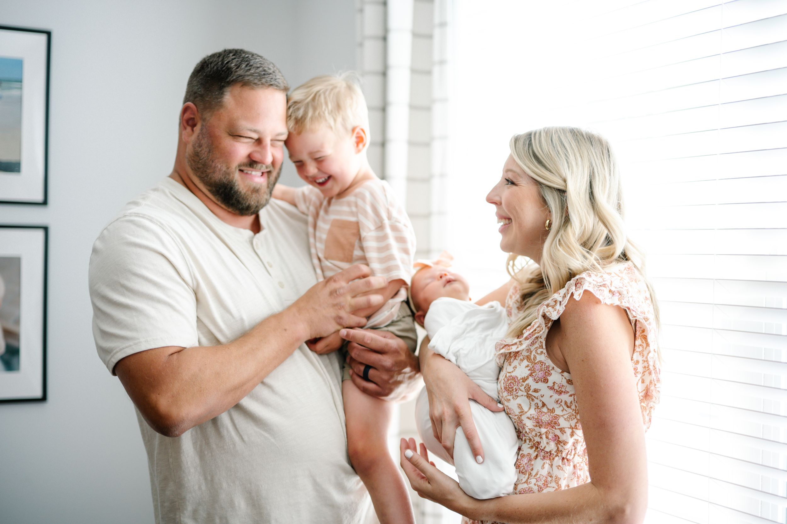 A happy family with a man and blonde woman smiling at their toddler and newborn baby near a bright window.
