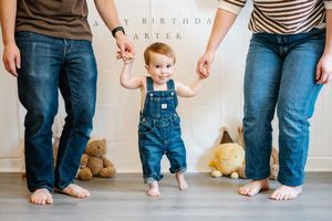 A baby in denim overalls takes first steps holding parents hands at a birthday party celebration.