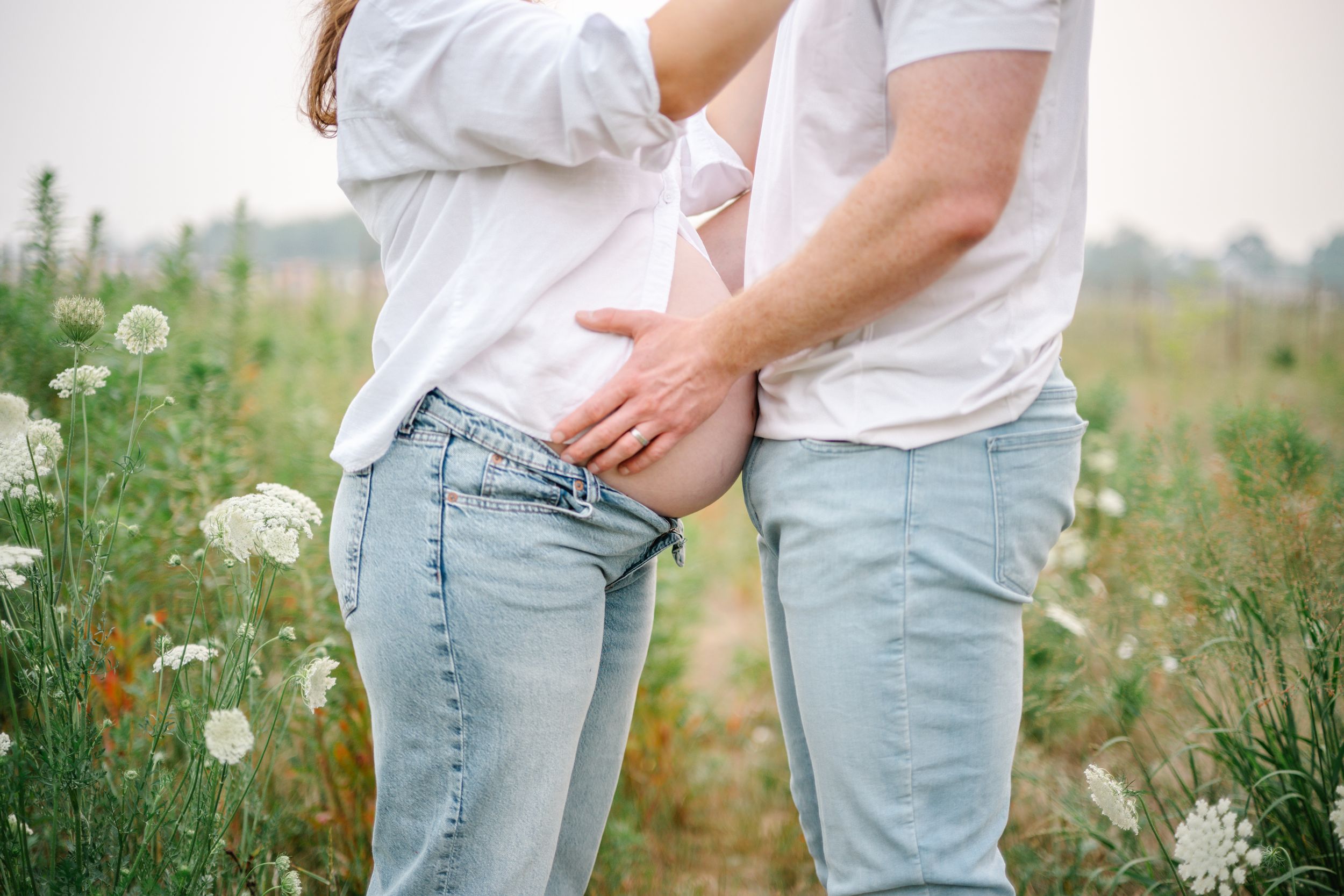Couple in white shirts and jeans embracing in a wildflower field, showing a pregnant belly outdoors.
