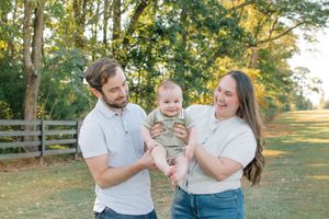 Happy young parents laugh while lifting their smiling baby together in a sunny outdoor park setting.