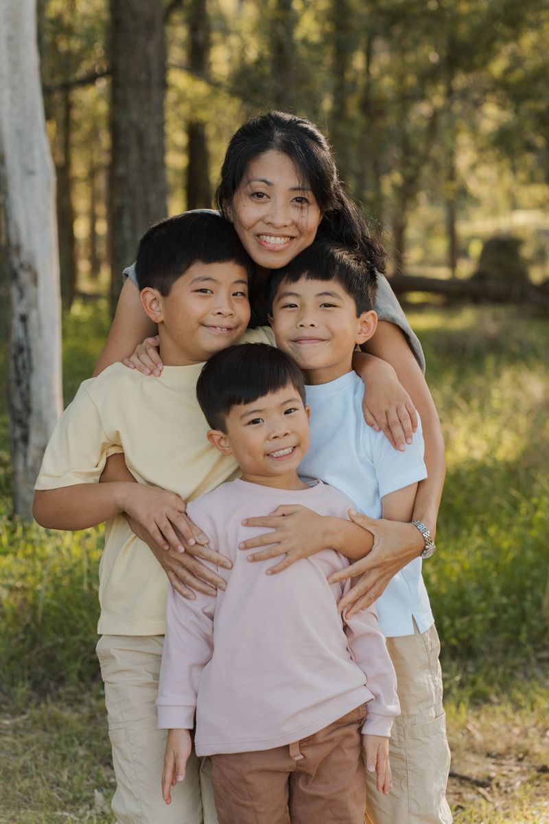 Smiling mother hugging three young sons outdoors in a sunny forest setting for a family portrait in rouse hill regional park