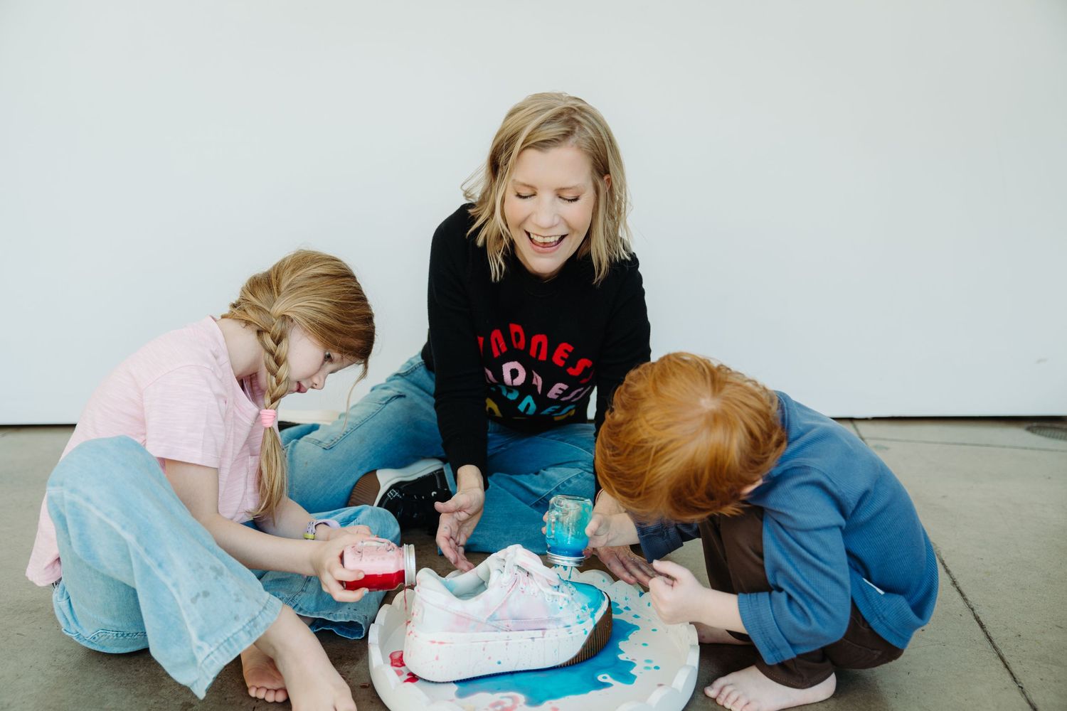 Woman and two children sitting on floor, laughing while painting white sneakers with colorful paint together.
