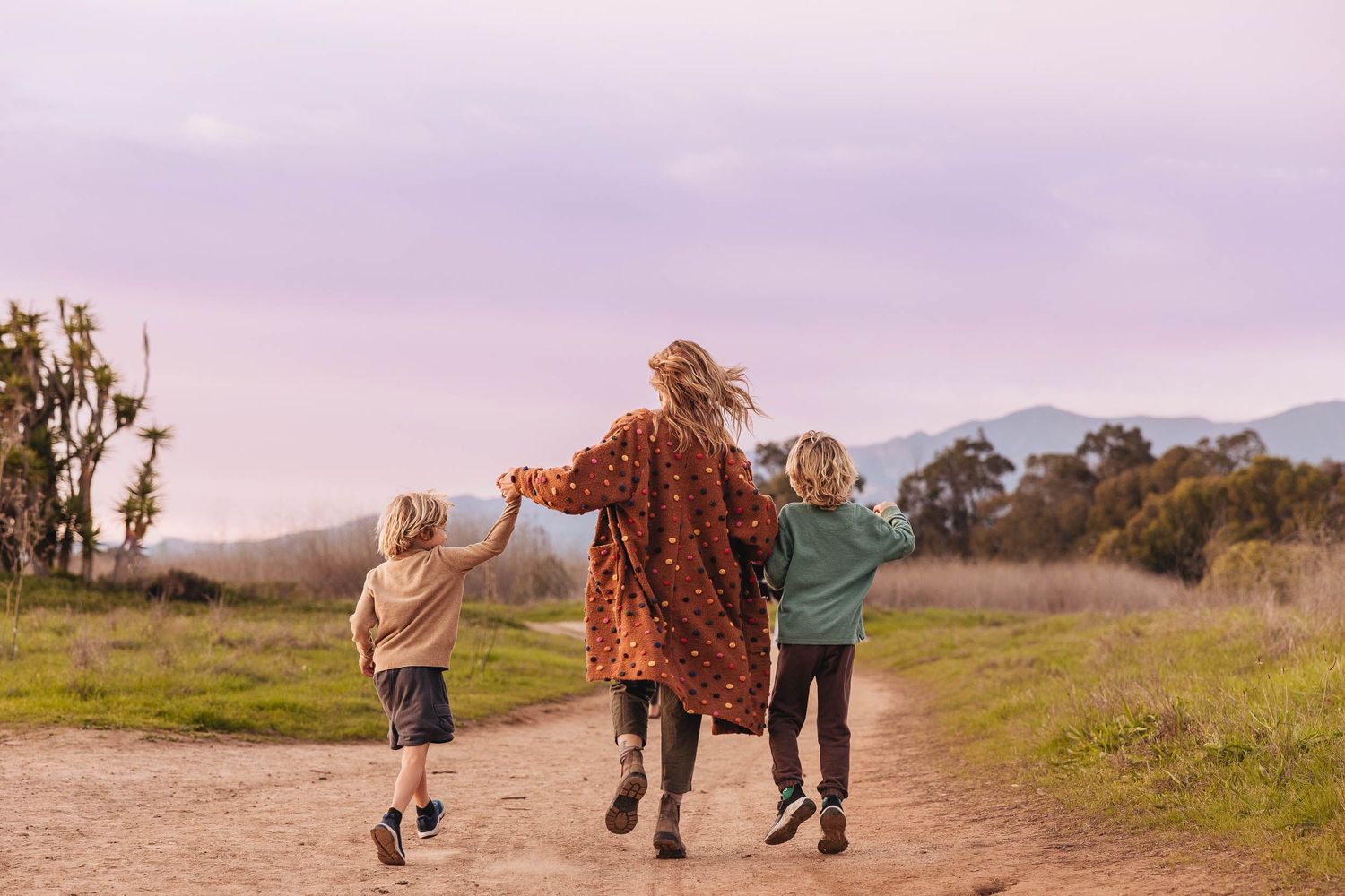 A woman and two children walk hand-in-hand down a dirt path through green fields under a purple sky.
