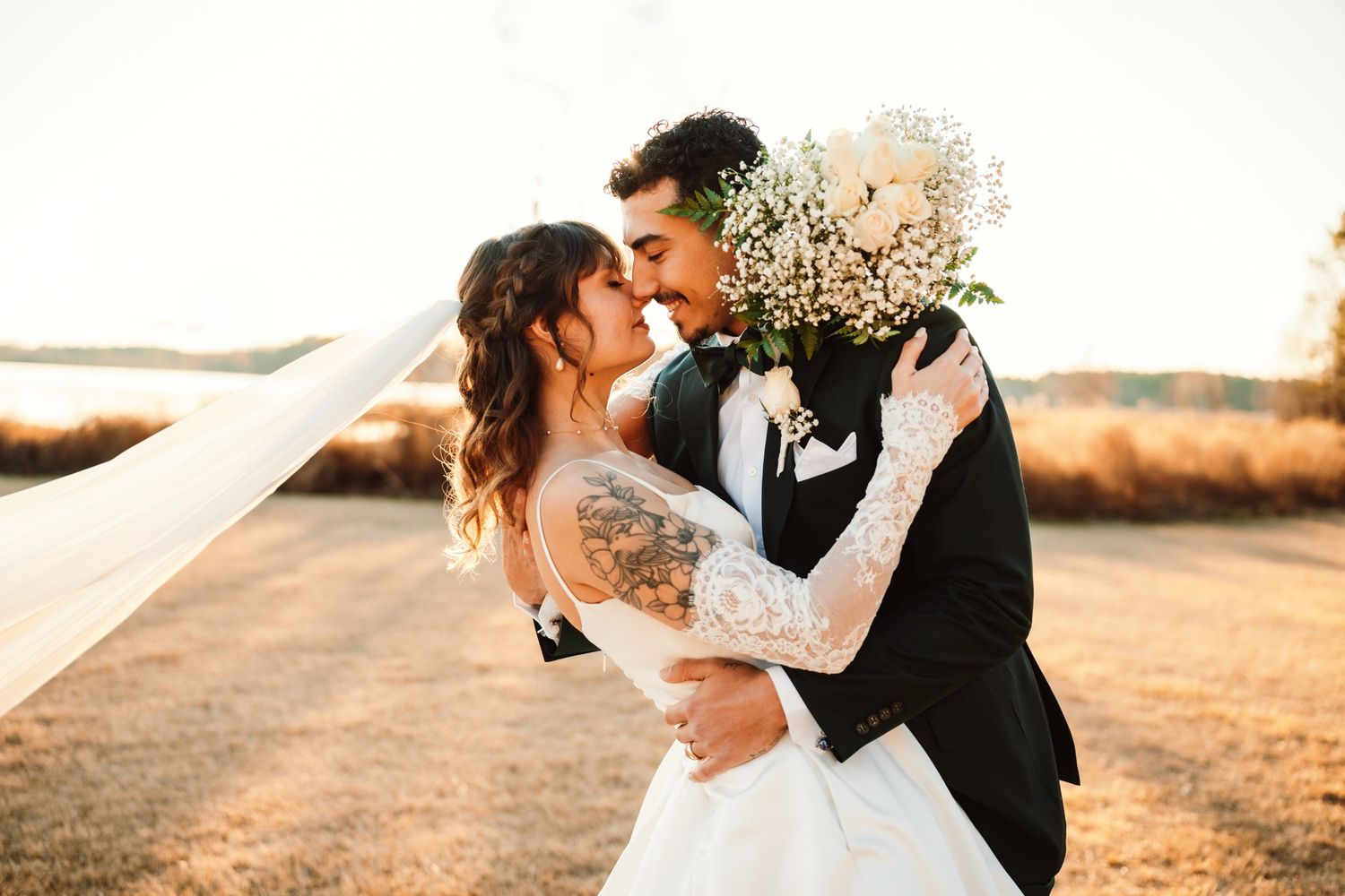 A bride and groom share a romantic kiss outdoors at golden hour, with a flowing veil and white bouquet.
