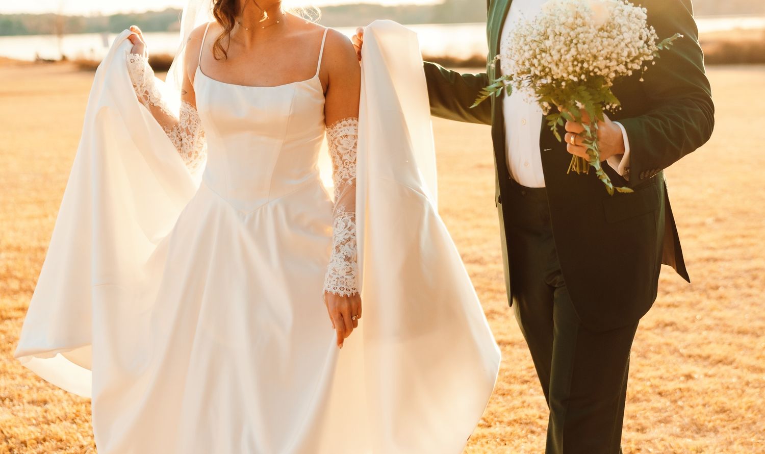 Bride in white gown holding veil walks beside groom in black tuxedo carrying bouquet outdoors.