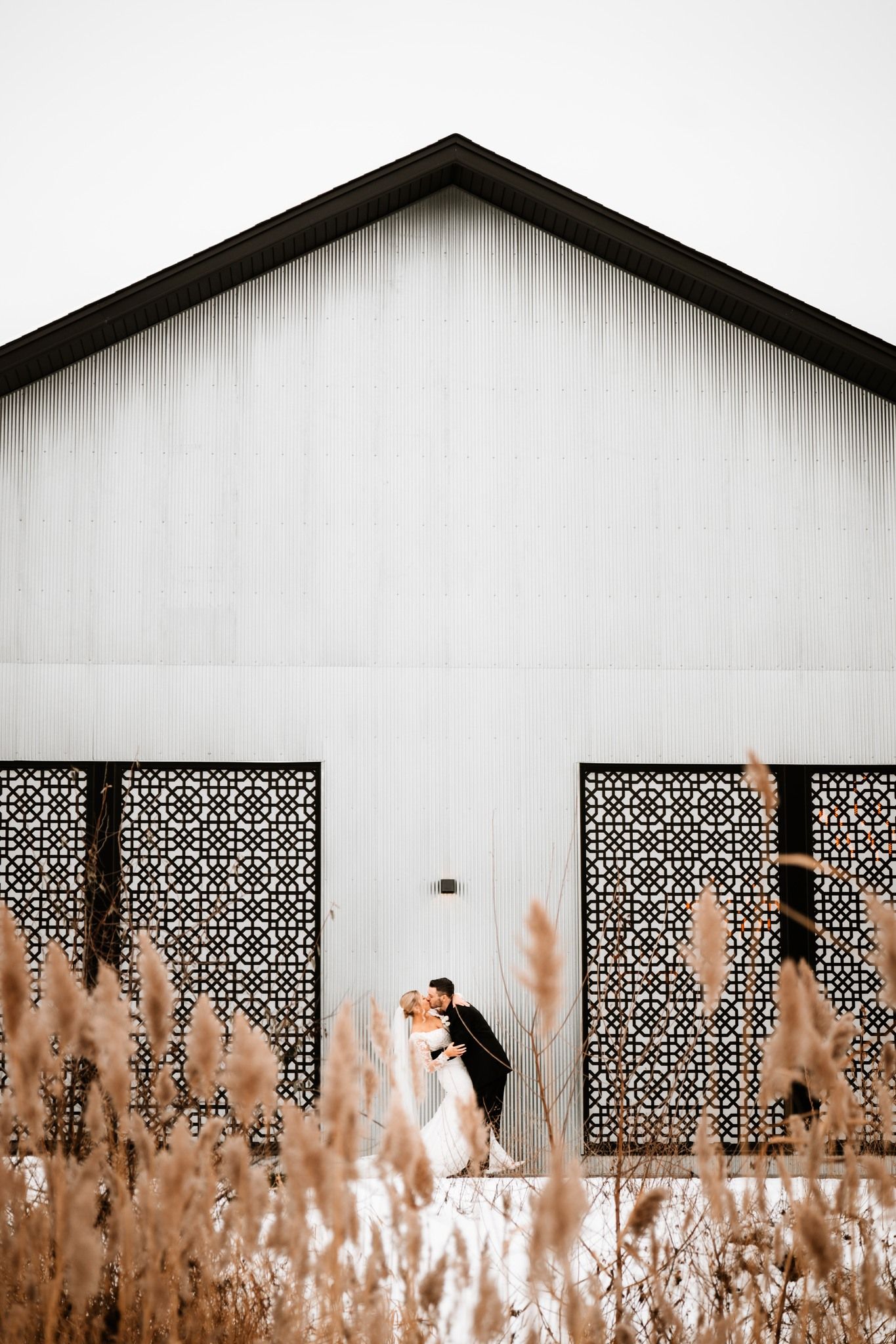 Bride and groom kissing in front of a white barn with decorative black panels and golden pampas grass in the foreground.