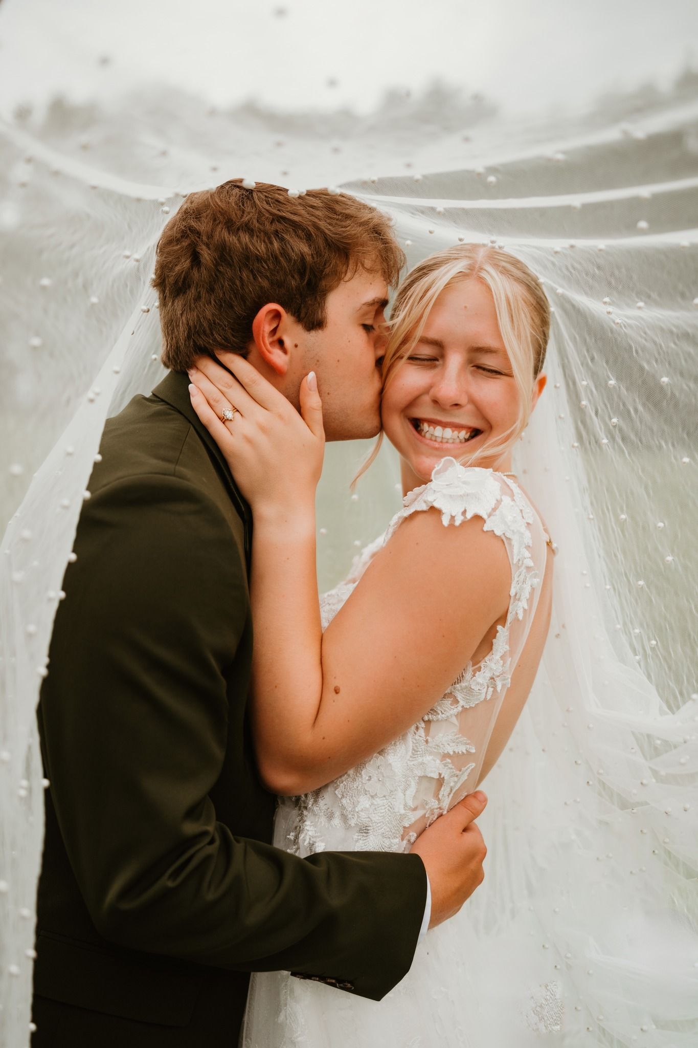 Bride laughing joyfully as groom kisses her cheek under a pearl-embellished wedding veil.