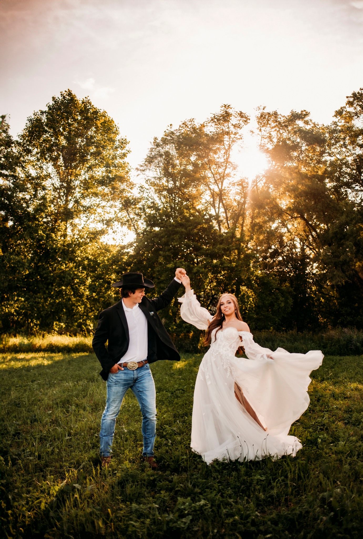 Bride and groom dancing in golden sunset light in a green meadow, bride twirling in flowing white gown.