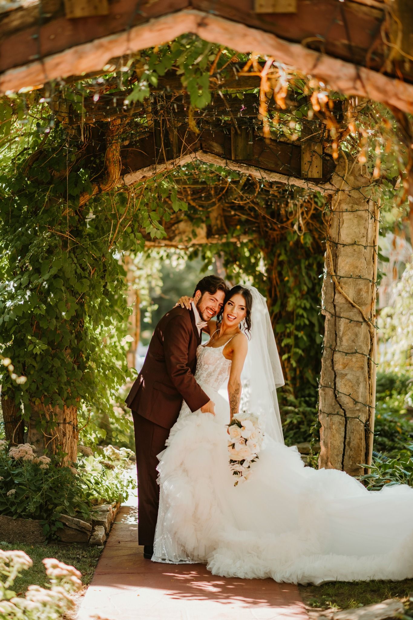 Bride and groom embrace under a lush garden arbor, bride in ruffled white gown holding bouquet, groom in brown suit.