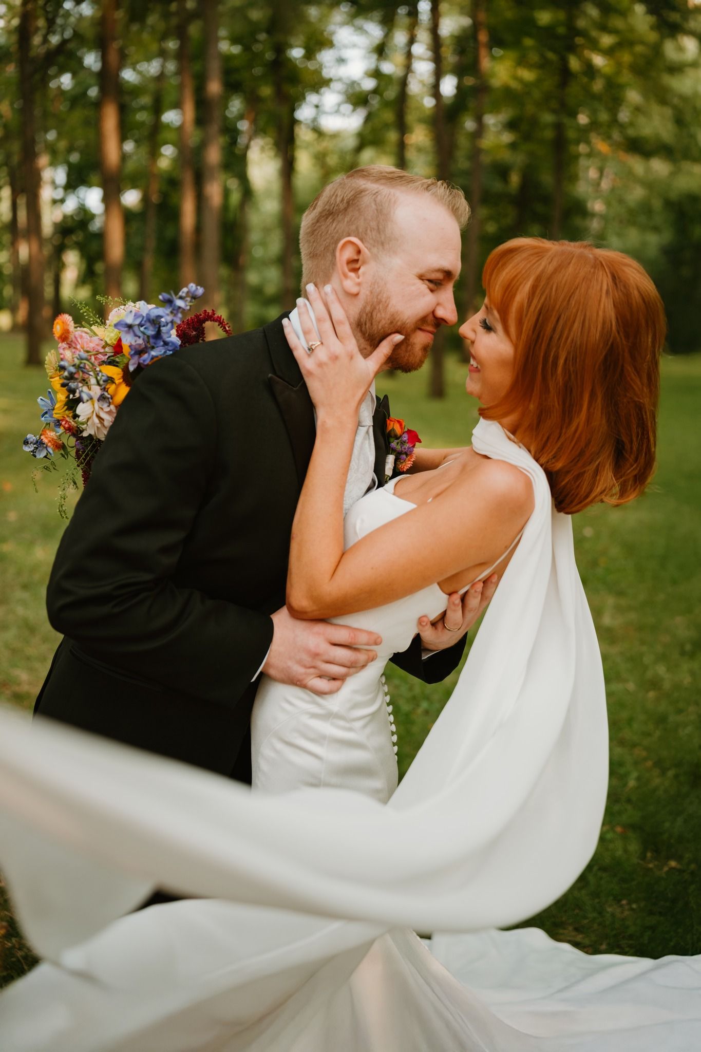 A bride and groom share a romantic moment outdoors, with her white gown flowing and a colorful bouquet visible.