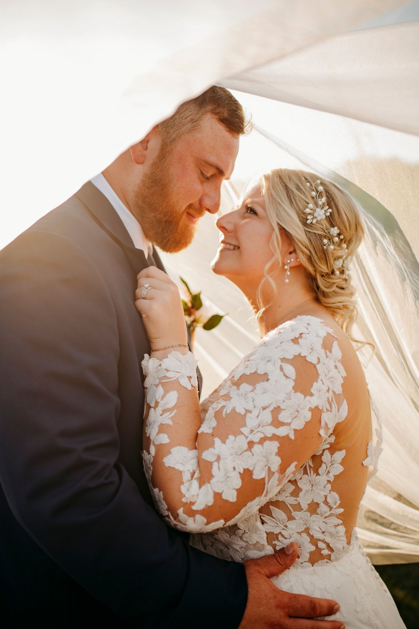 Bride and groom share a romantic moment under a veil in golden sunlight, bride wearing lace gown with floral headpiece.
