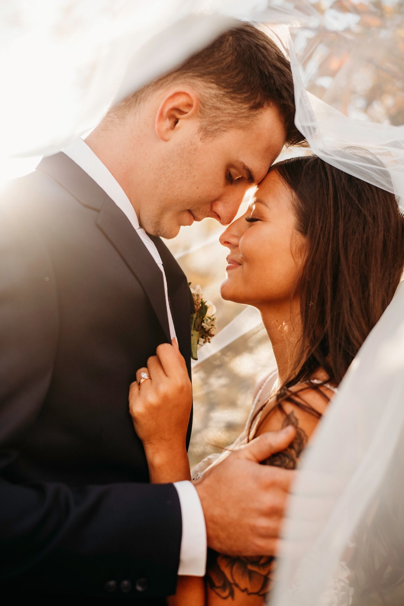 Bride and groom sharing an intimate moment under a veil in warm sunlight, foreheads touching tenderly.
