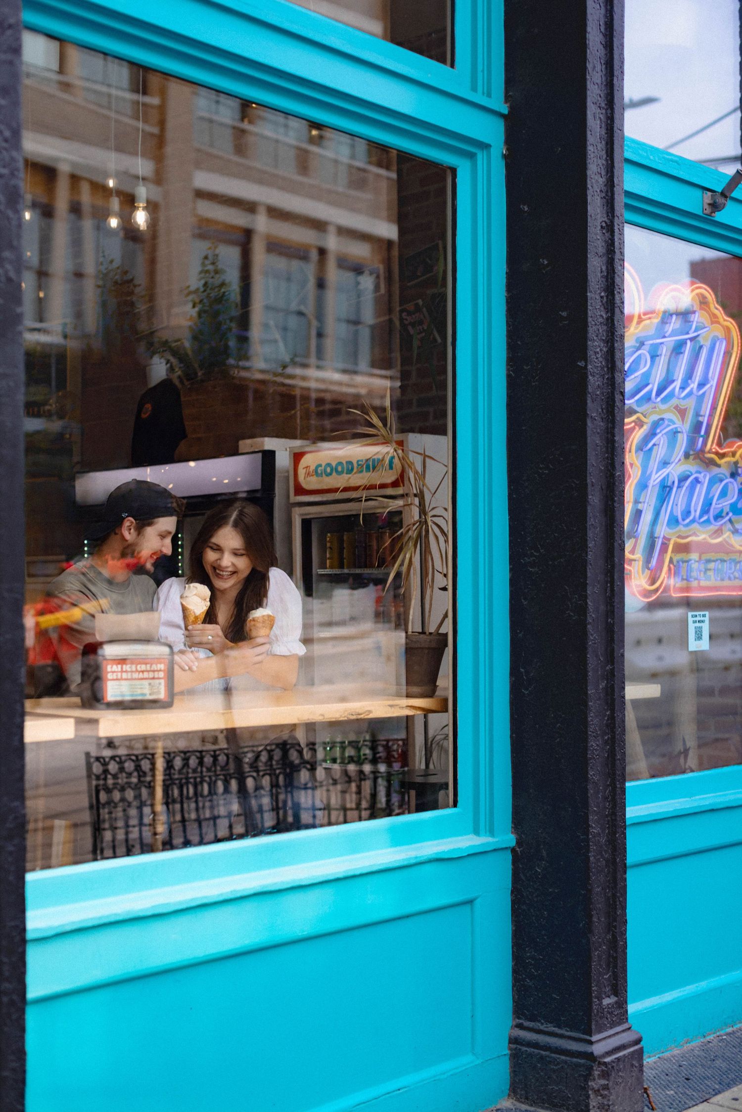 A man and woman sit at a counter through a window inside a bright blue building eating ice cream with a neon Betty Rae's sign next to them.