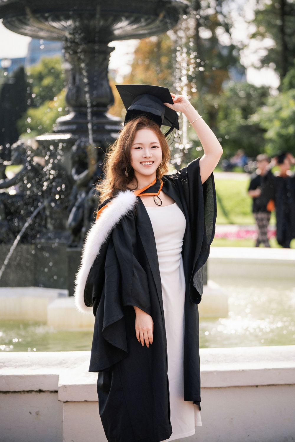 Joyful graduate raising her cap in front of the landmark fountain at Albert Park for celebratory graduation photos.