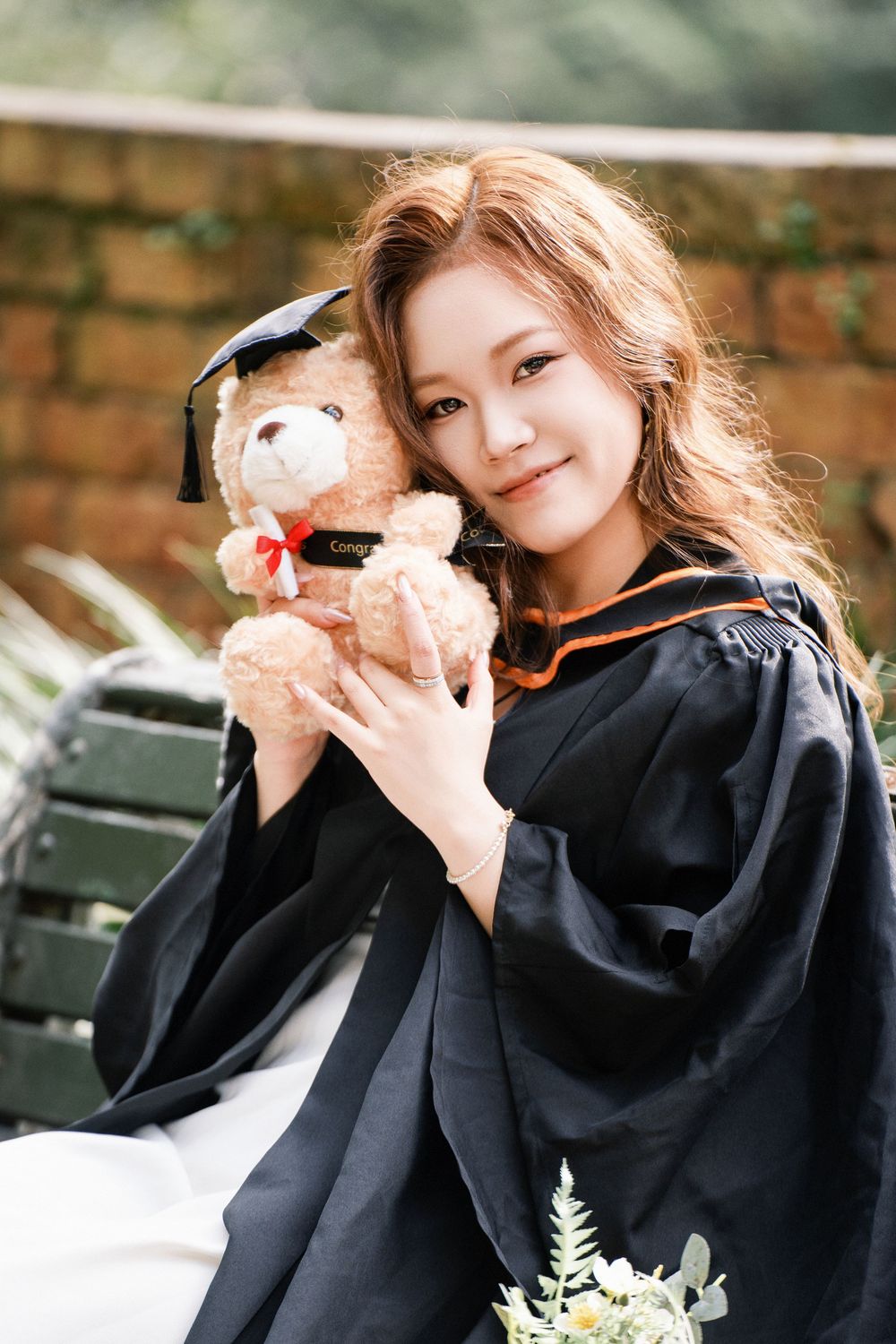 Happy student holding a graduation bear against a classic stone wall at Albert Park, Auckland.