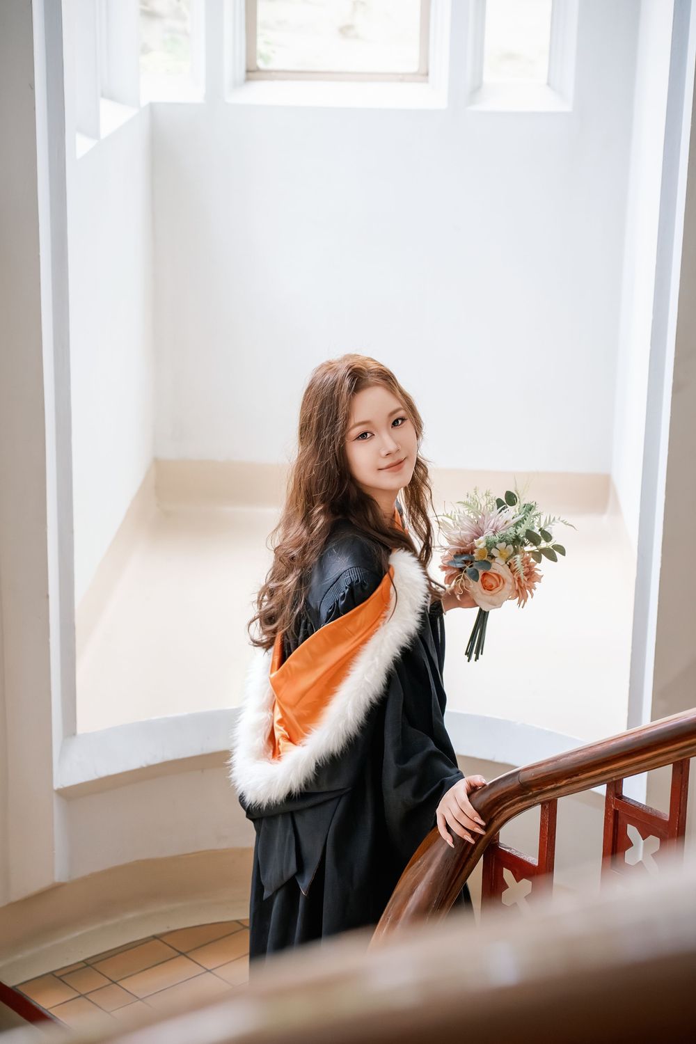 Graduate holding a bouquet while walking down the elegant wooden staircase inside the UoA Clock Tower.