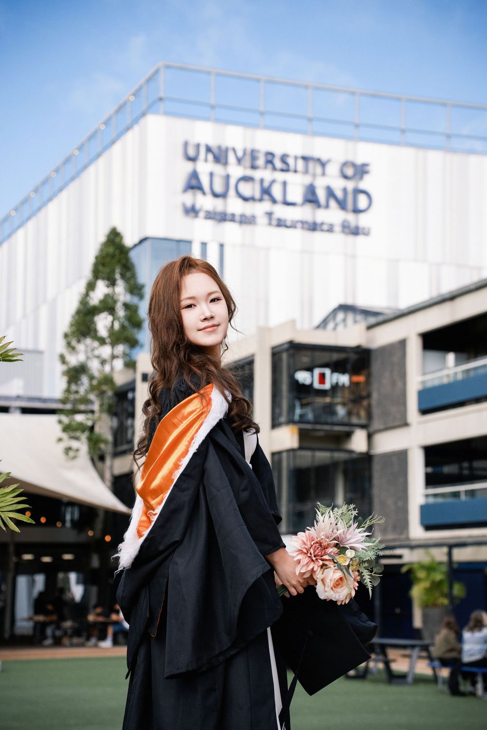 Proud graduate in academic dress posing outside the University of Auckland (UoA) campus.