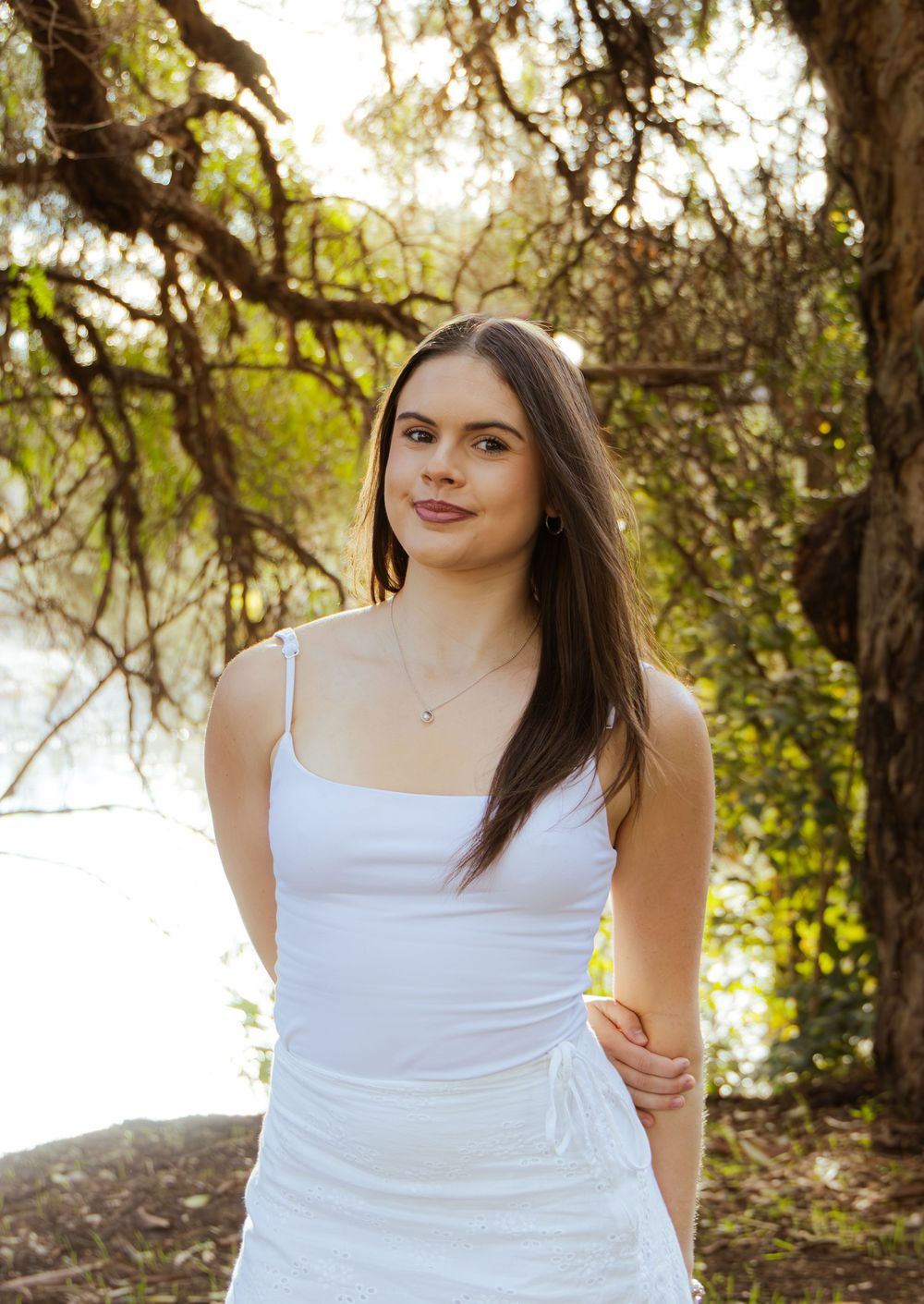 Young brunette woman in white sundress standing under trees near water in golden sunlight.