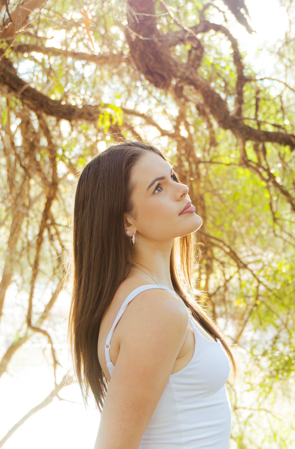 Brunette woman in white tank top looking up through sunlit tree branches in a natural setting.