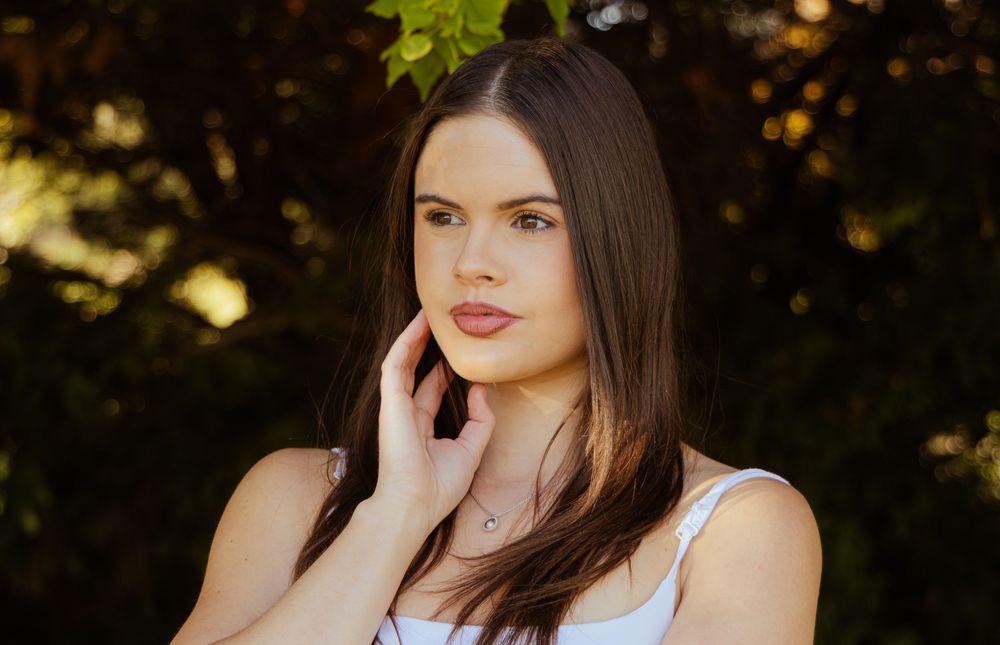 Close-up portrait of a young brunette woman in white top touching her face among green foliage.