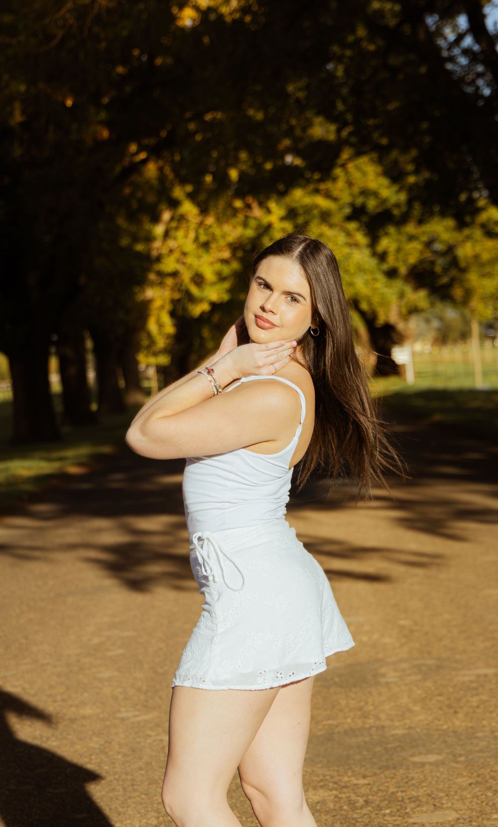 Young woman in white mini dress posing outdoors among autumn trees in warm golden light.