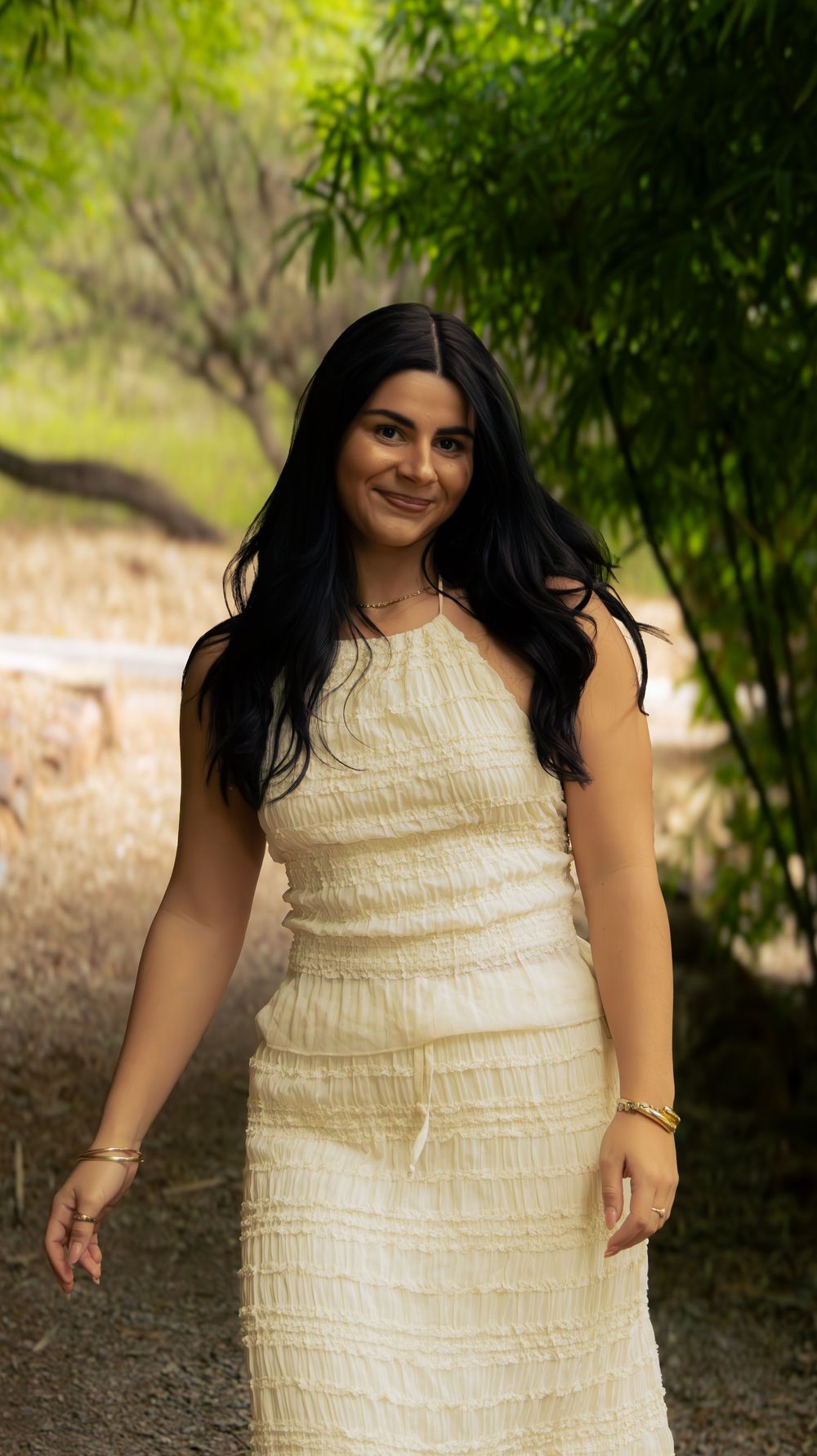 Young woman with long black hair in cream ruched dress smiling outdoors among green trees.