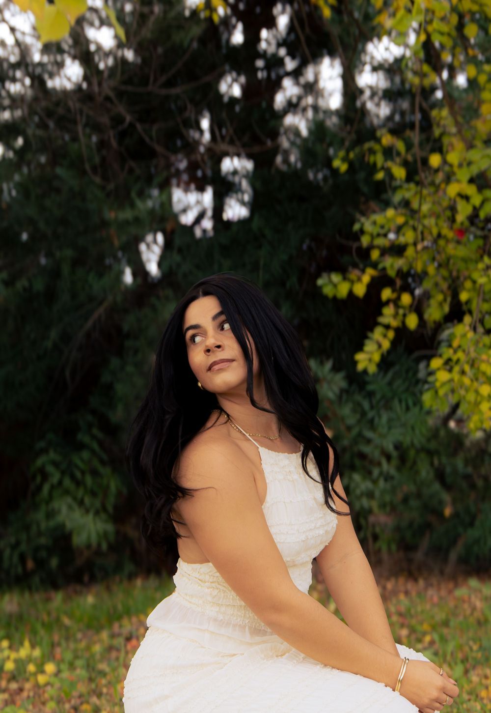 Young woman with long black hair in white dress sitting outdoors looking up among autumn trees.