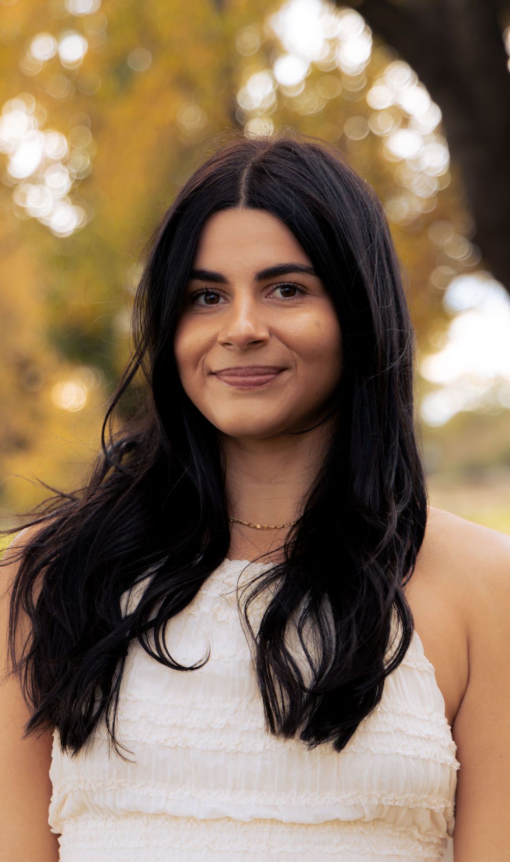 Young woman with long black hair smiling in white sleeveless top with autumn bokeh background.