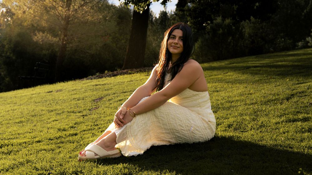 Young woman in cream dress sitting on green grass in golden sunset light under a tree.