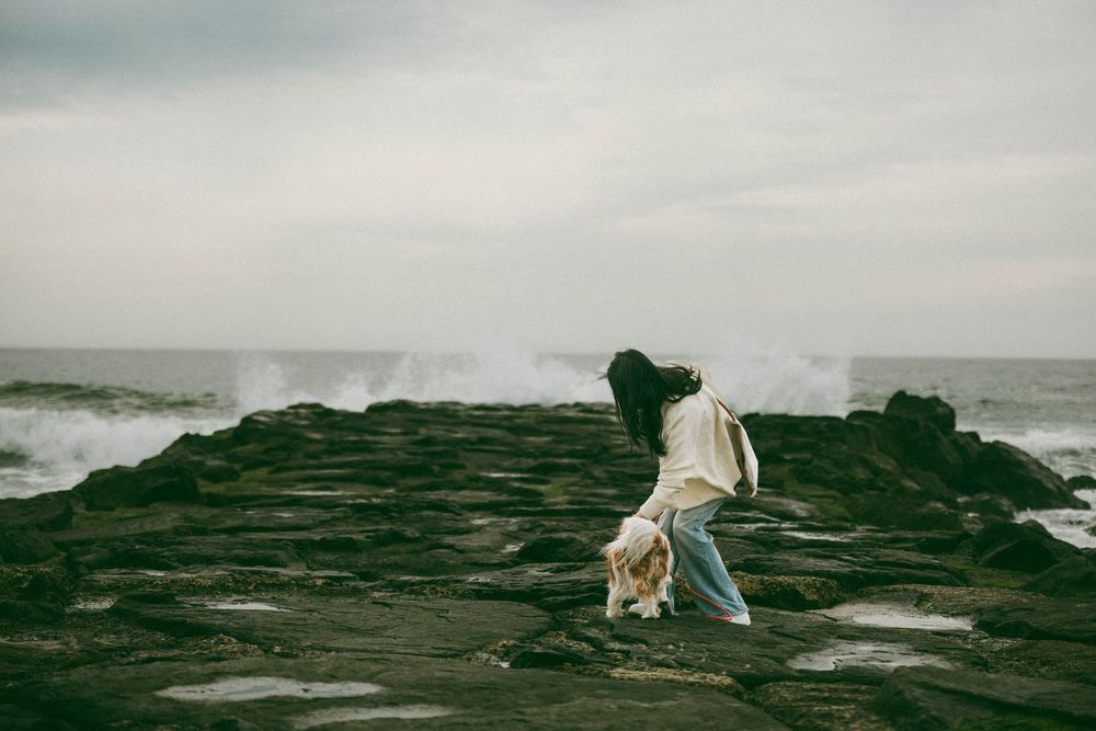 Person walking on rocky coastal cliffs with dramatic ocean waves crashing in the background on an overcast day.