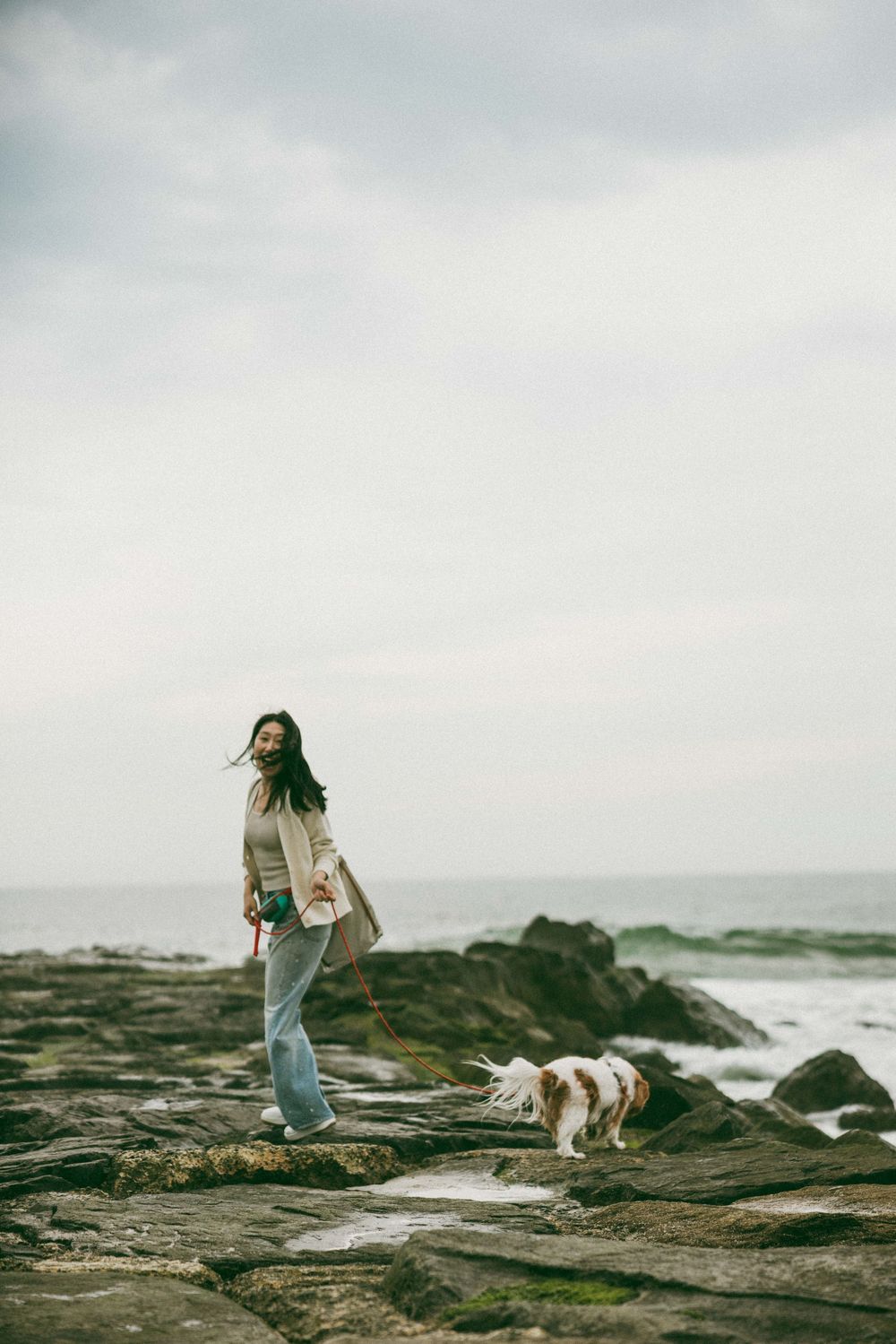 Woman walking her Cavalier King Charles Spaniel on rocky coastal shoreline on an overcast day.
