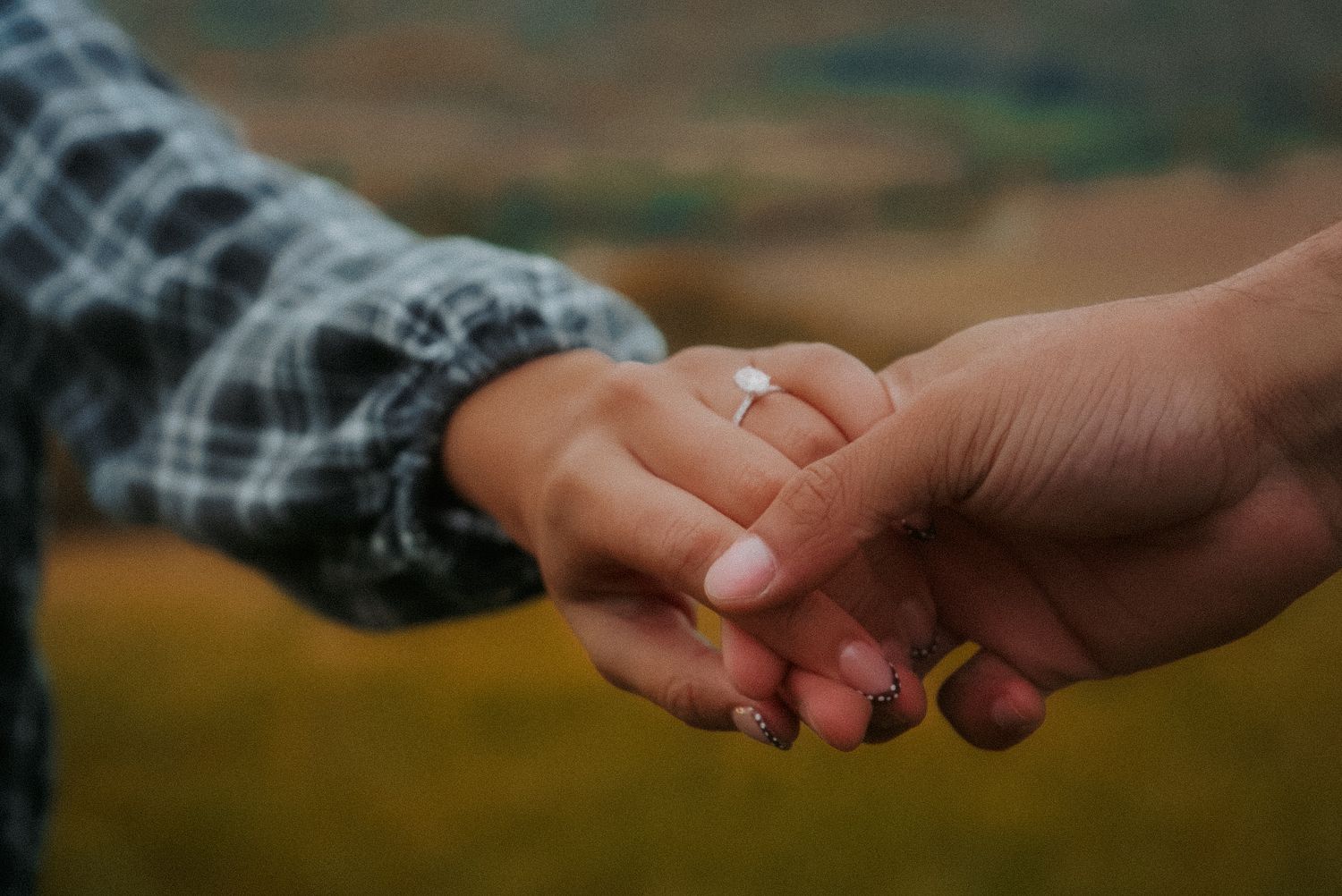 Close-up of couple holding hands with engagement ring on woman's finger in romantic outdoor setting.