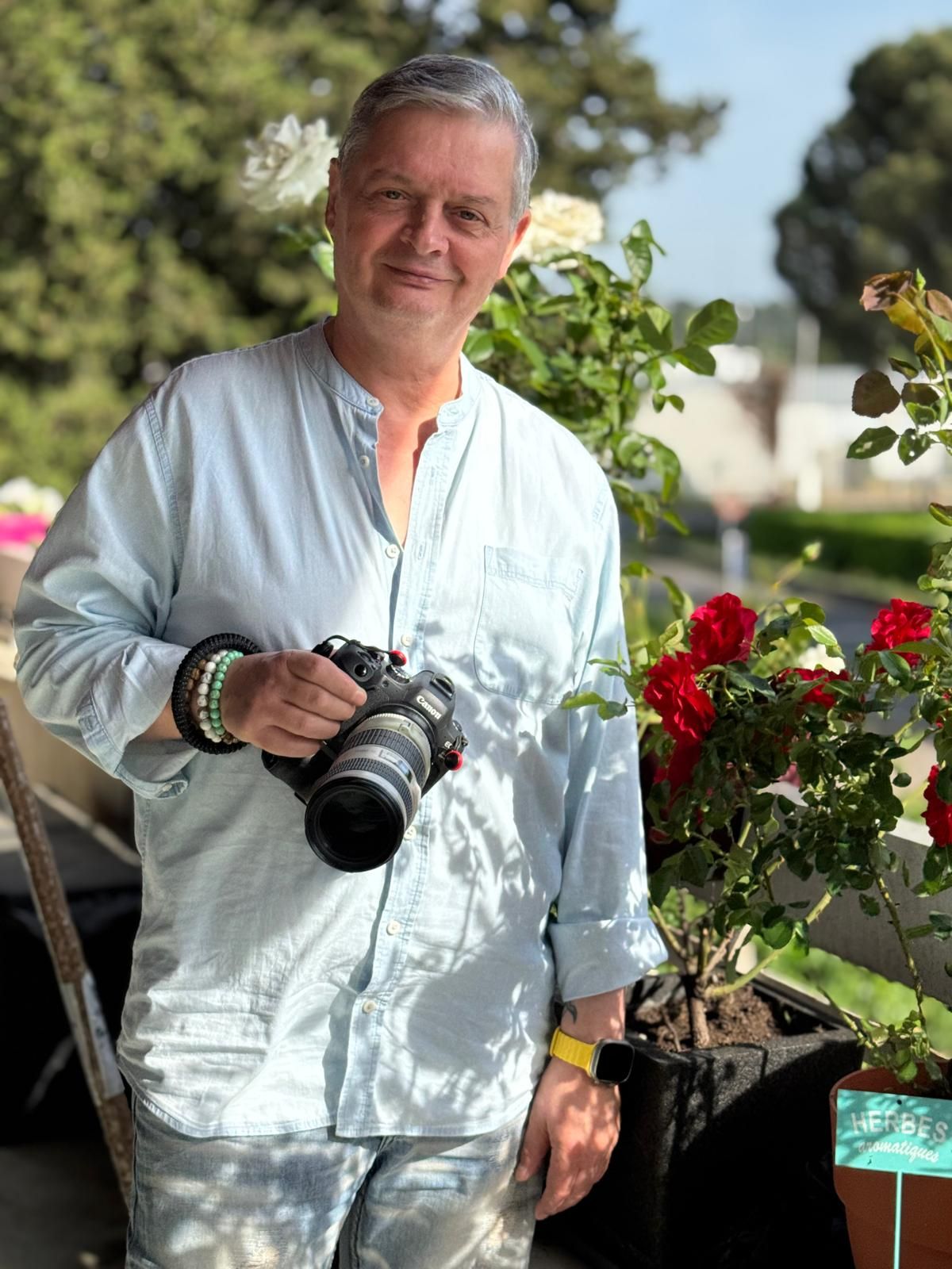 Smiling man holding a professional camera standing beside red and white rose plants outdoors.