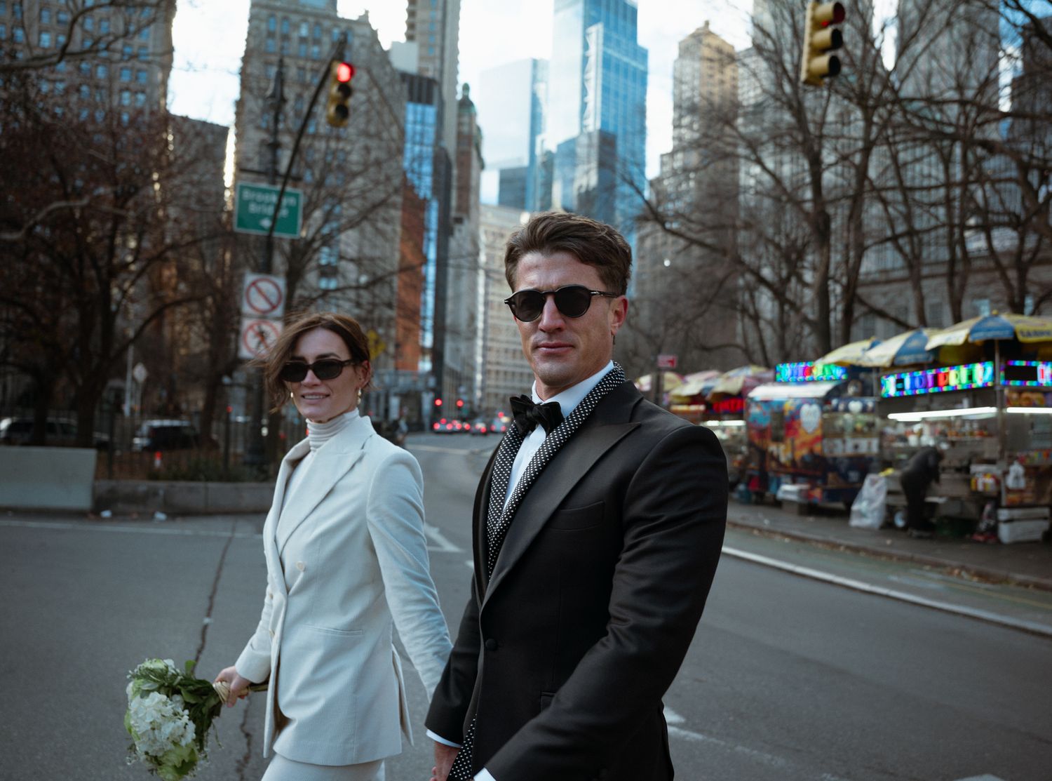 Elegant couple in wedding attire posing on a New York City street, woman in white suit holding bouquet, man in black tuxedo.
