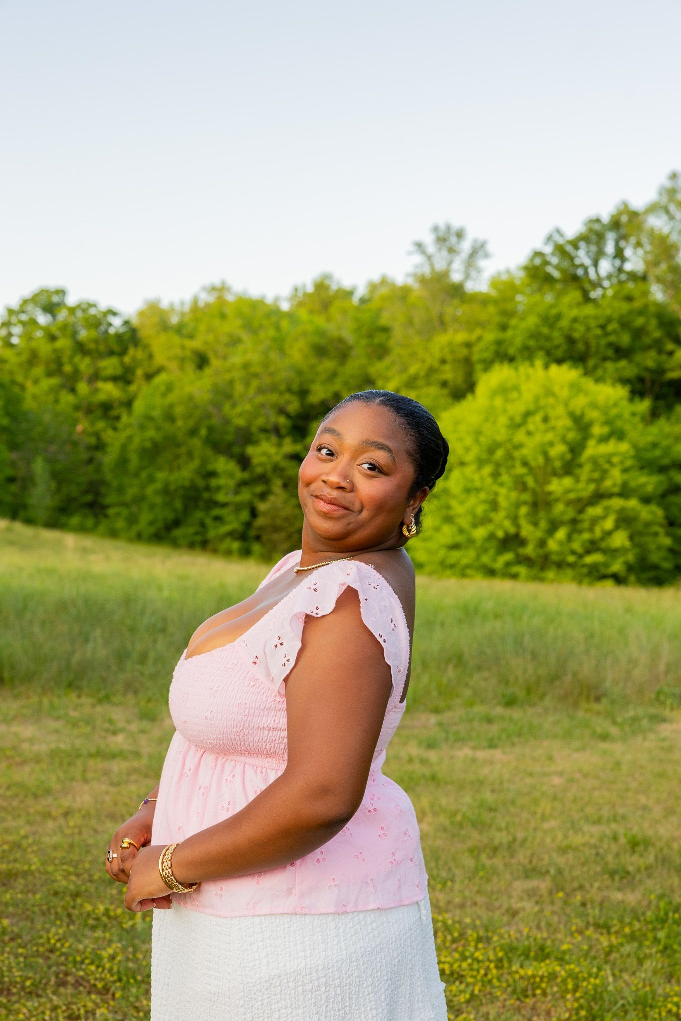 Young Black woman in pink top and white skirt smiles outdoors in green field at golden hour.