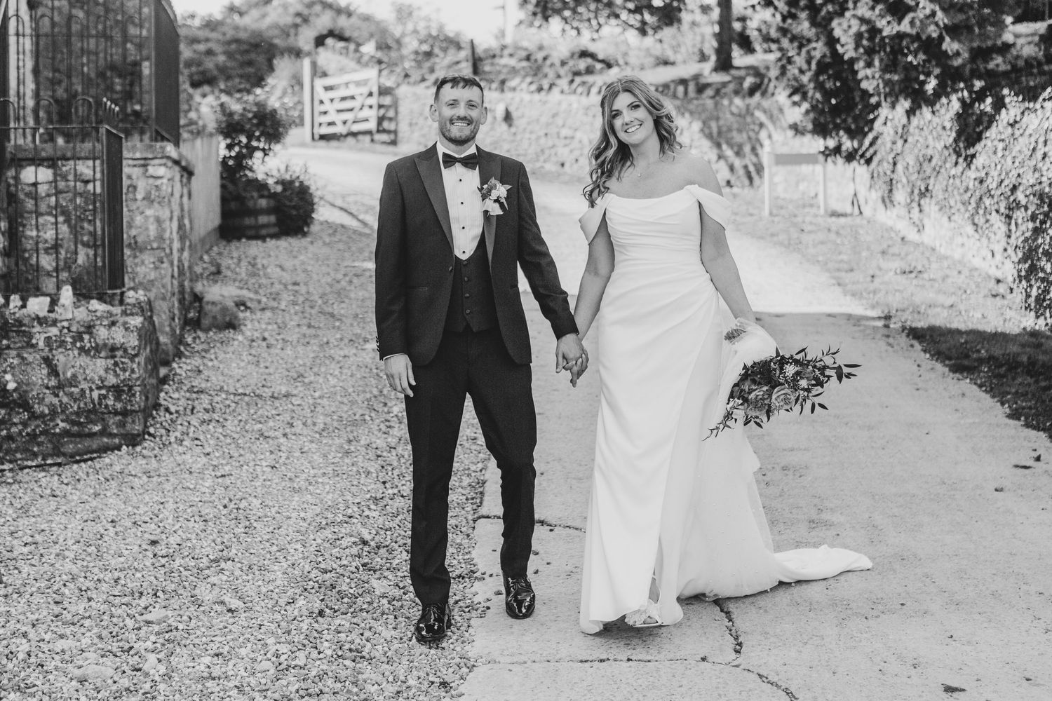 Black and white photo of a smiling bride and groom holding hands while walking along a gravel path on their wedding day.