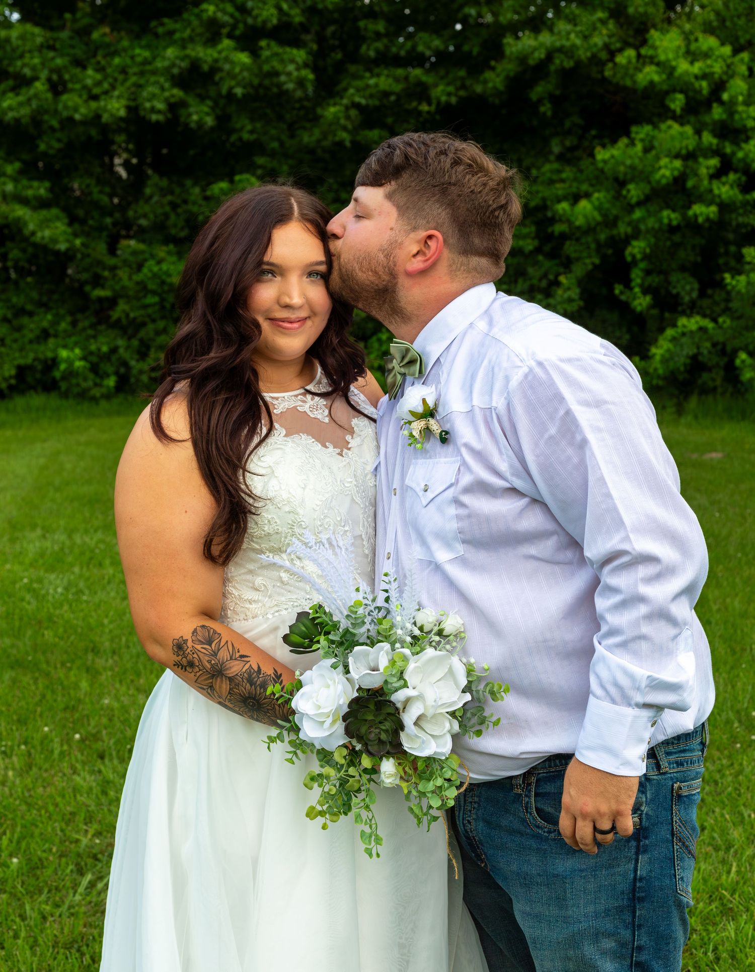 Groom kissing bride on cheek outdoors, bride holding white floral bouquet in wedding dress with lace bodice.