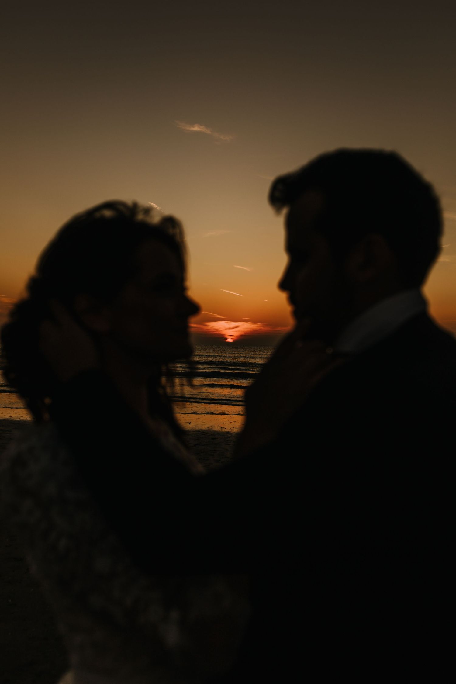 Silhouette of bride and groom facing each other on beach at stunning orange sunset.