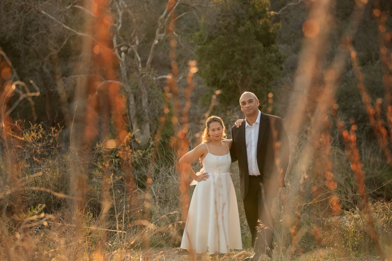 Couple in wedding attire standing together in golden sunlit bushland during outdoor photo session.