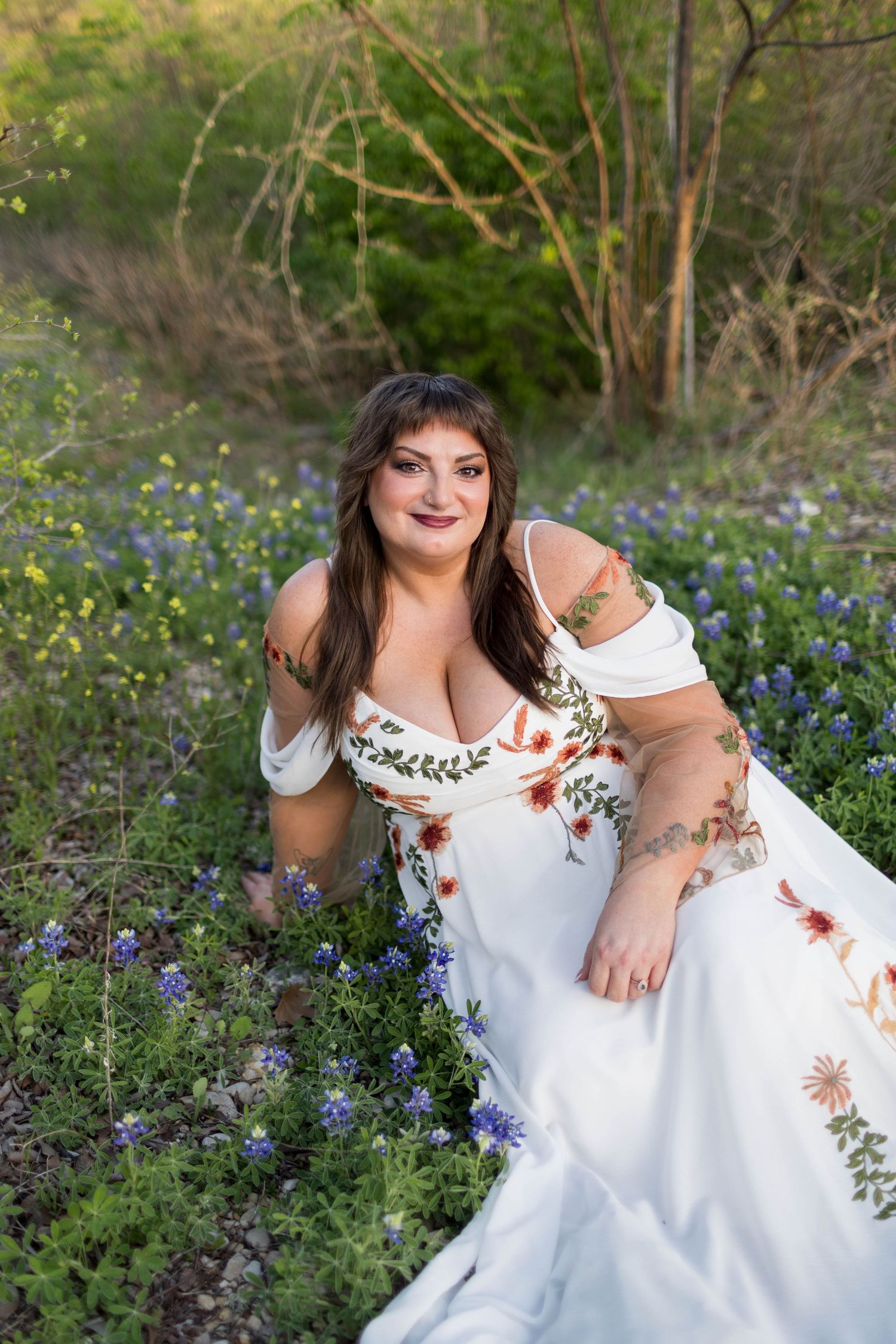 Woman in floral white off-shoulder gown sitting among bluebonnets in a wildflower field, smiling warmly at McKinney Falls State Park.
