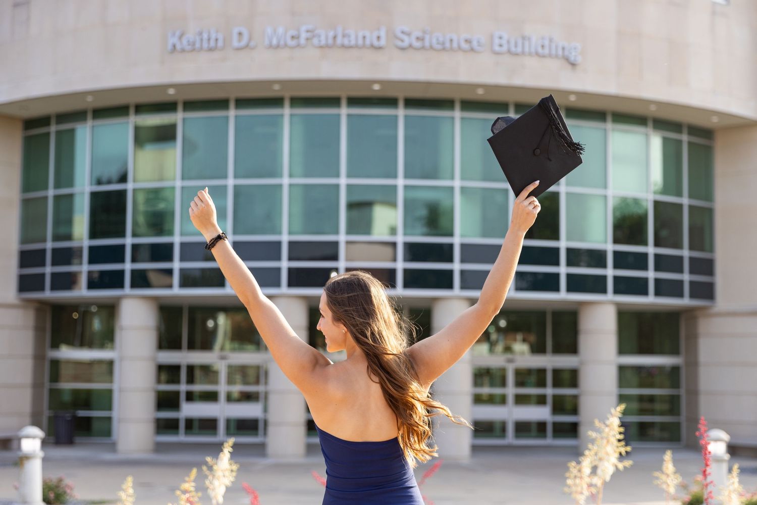 picture of a girl, hands raised in achievement holding her graduation cap standing in front of the college science building