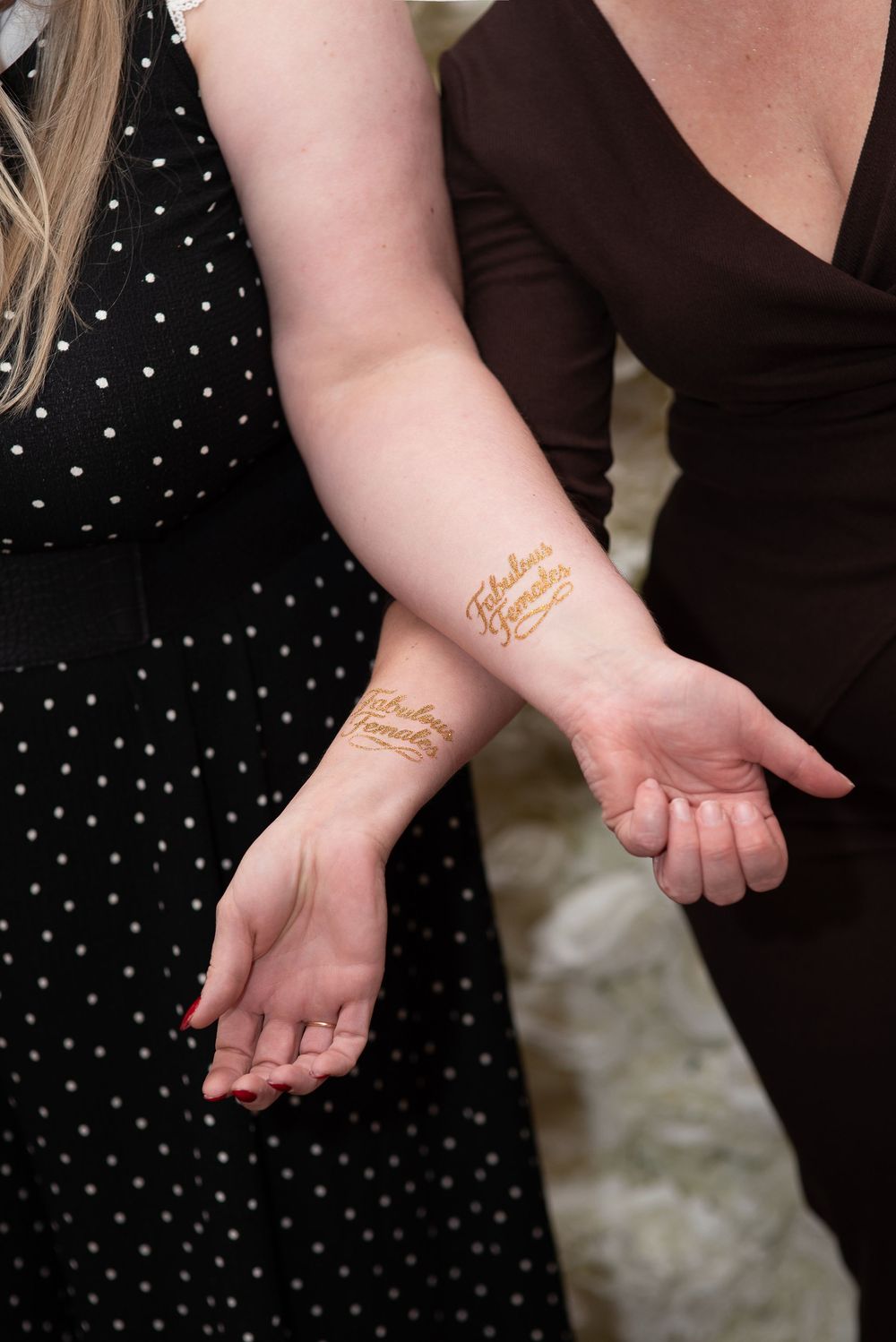 Two women showing matching gold metallic temporary tattoos reading 'Fabulous' on their wrists and forearms.