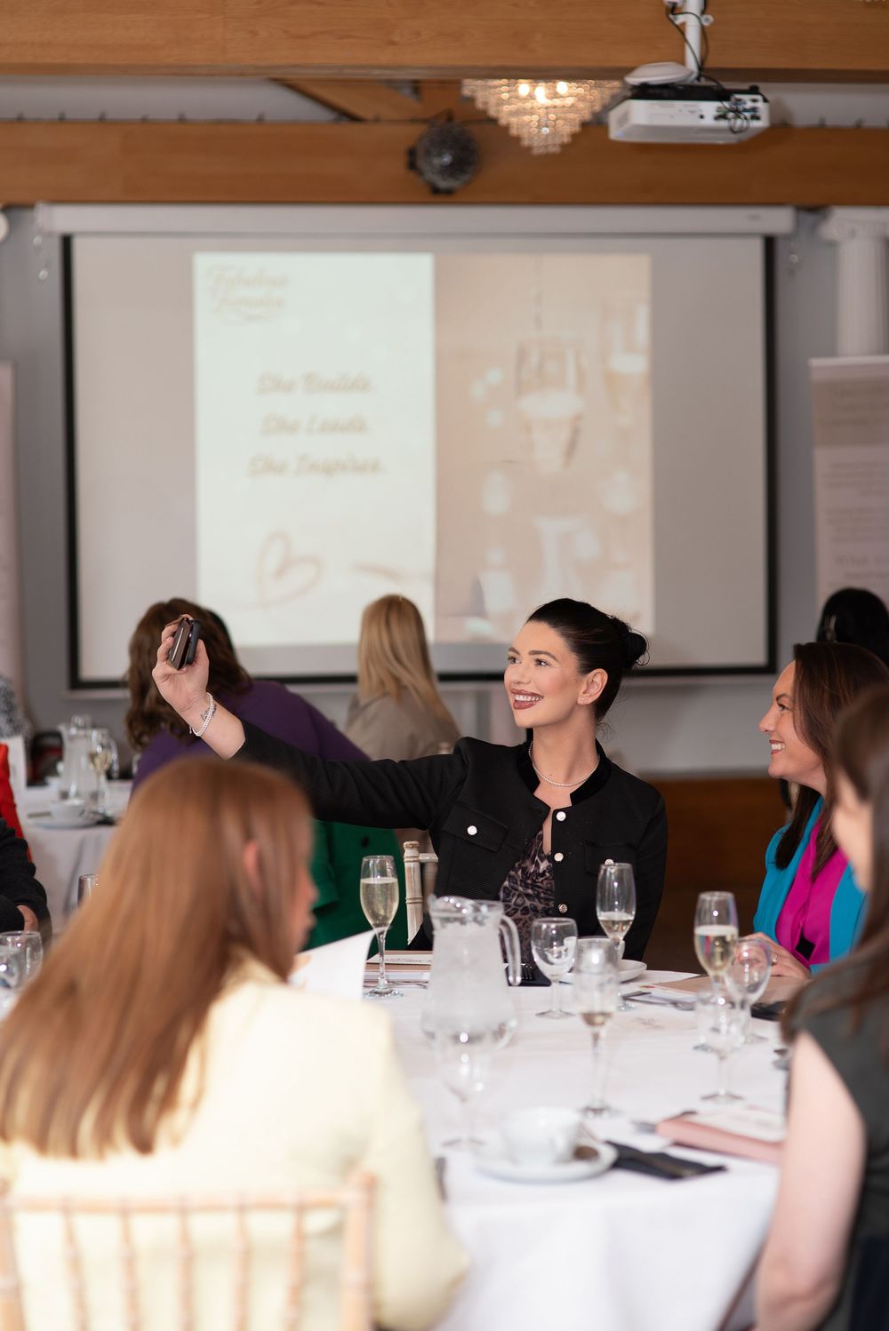 Woman taking selfie at elegant event with projector screen, wine glasses, and smiling guests at white tablecloth tables.